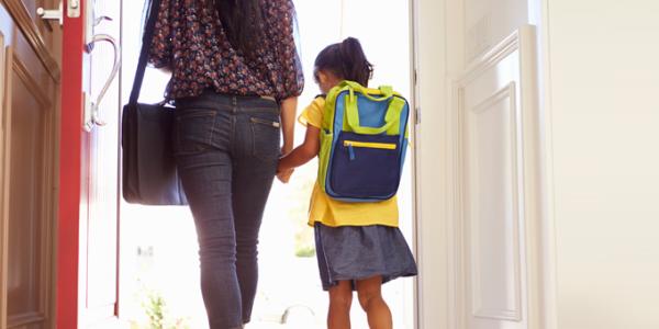 A young girl with a backpack walks through a door with her parent. Both are seen from behind.